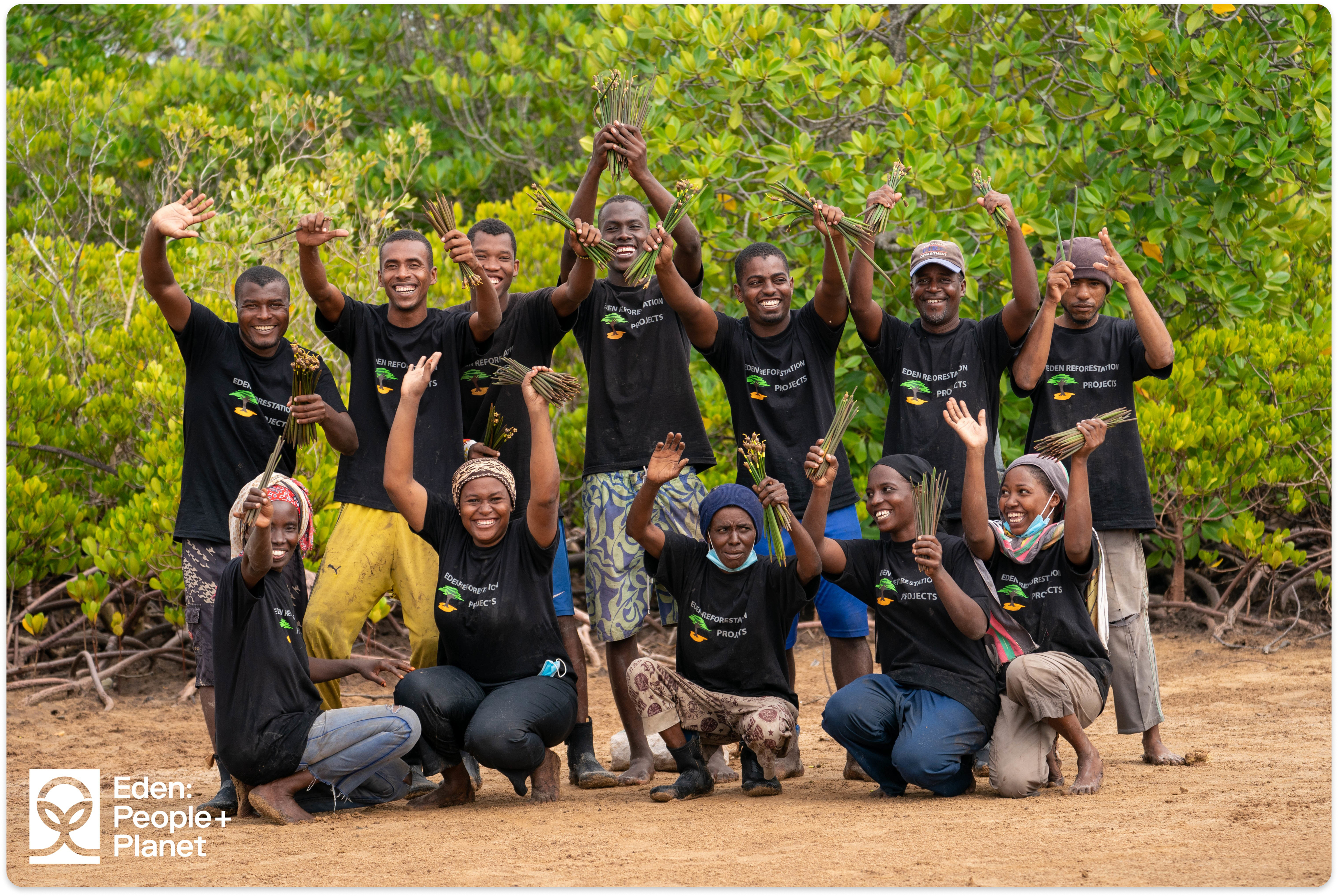 A group of Eden: People+Planet team members smiling and raising mangrove seeds in a forest clearing.