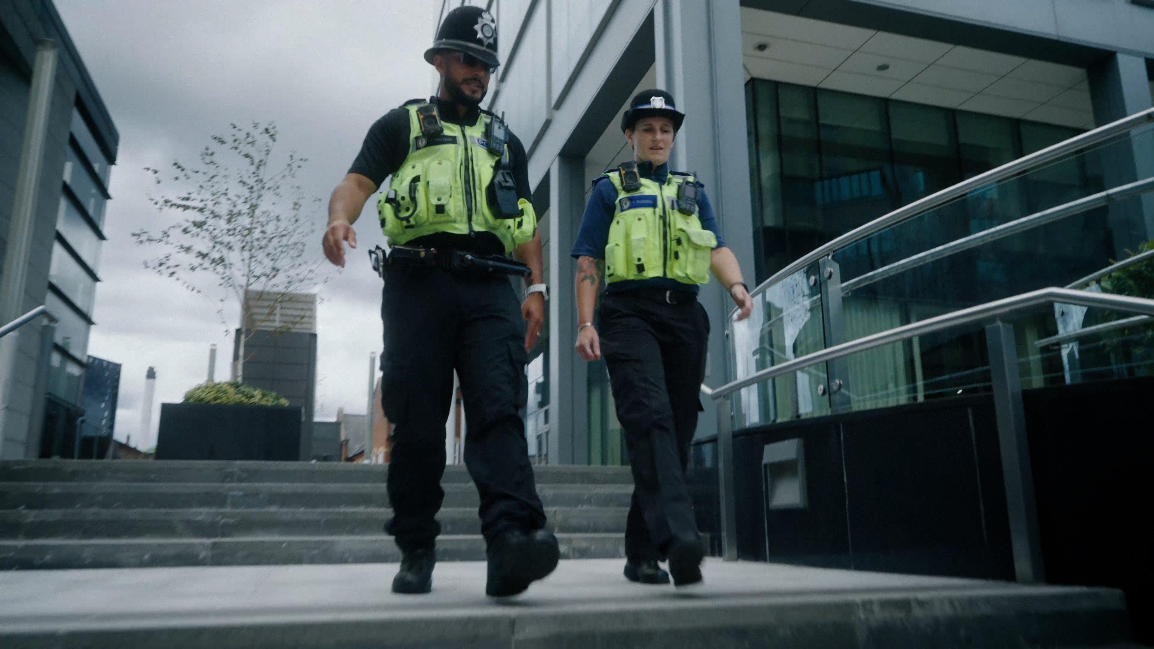 Two police officers in high-visibility uniforms walking down an outdoor staircase beside a modern office building.